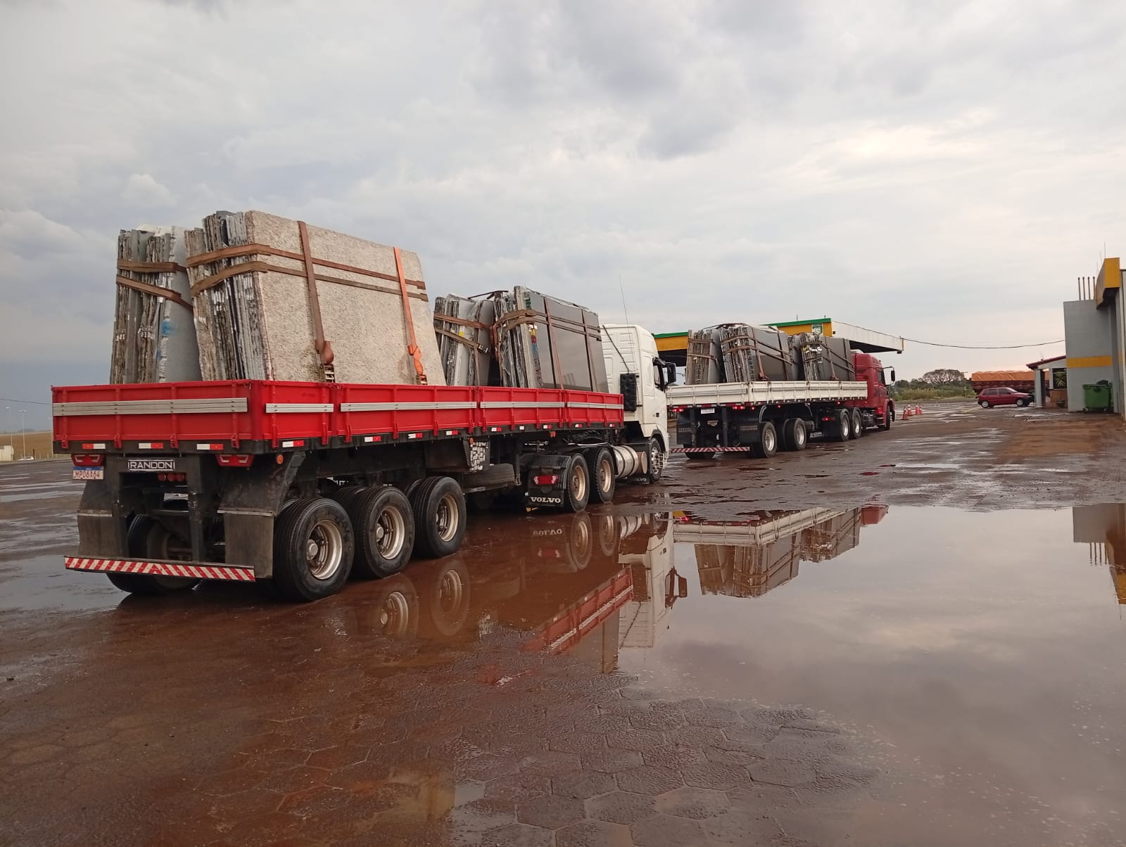 Duas carretas da TR Maizena carregadas de chapas de granito no pátio molhado, com o reflexo dos conjuntos na poça